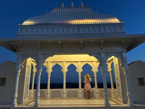 Girl stands beneath an illuminated ivory pavilion decorated with arches and domes under the evening sky.