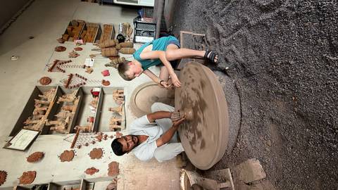 Potter guides a child shaping clay on a large wheel inside a rustic workshop filled with terracotta crafts.