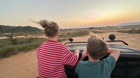 Two children ride standing in an open jeep across sandy terrain at sunset with hair blowing in the wind.