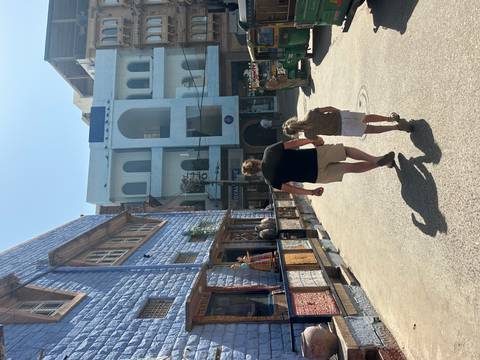 Parent and child stroll through a sunlit narrow street lined with blue-painted architecture and shops.