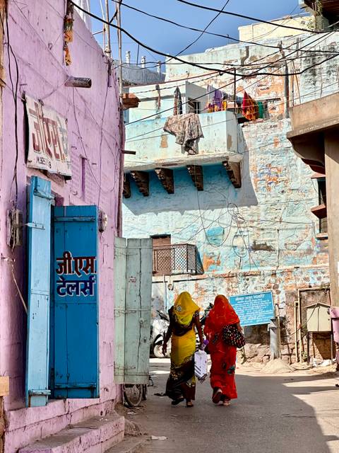 Vibrant alley of pastel walls where two sari-clad women walk past rustic balconies and signage.