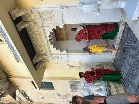 Women in red shawls greet a young girl at a carved marble doorway offering a ceremonial tray.