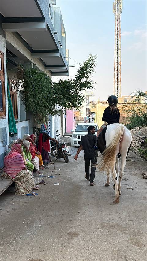 Girl on horseback is led through a village lane while older women sit chatting nearby.