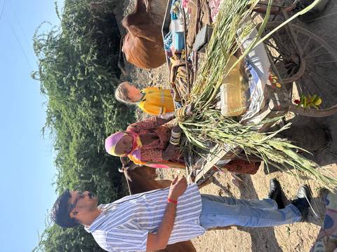 Local woman and girl prepare animal fodder at a rustic cart while a man looks on, with cows in the background.