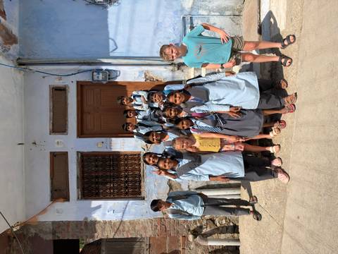 Group of local schoolchildren pose smiling with two visiting kids outside a blue-painted building.
