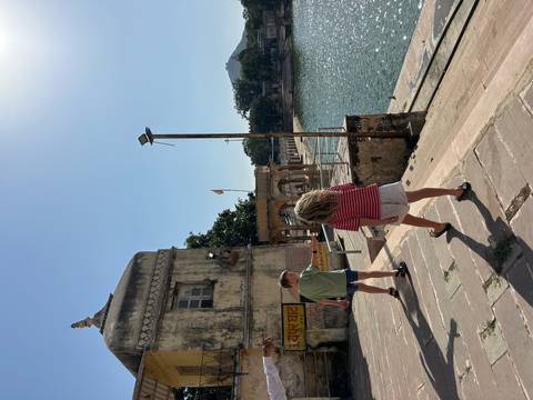 Two children wander along the stone steps of a lakeside ghat beside turquoise water under a bright sky.