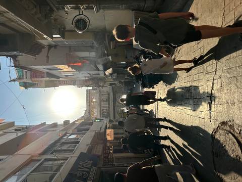 Backlit street scene with tourists and locals walking through a narrow alley of shops in the late afternoon sun.