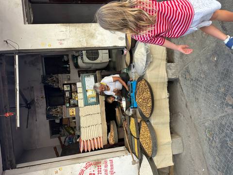 Market vendor sorts nuts in large trays inside a rustic shop while a child in a striped shirt looks on.