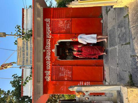 Two visitors enter a bright red Hanuman temple with Hindi script and decorative bells above the doorway.