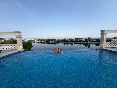 Two children enjoy an infinity rooftop pool overlooking a tranquil lake and cityscape on a bright day.
