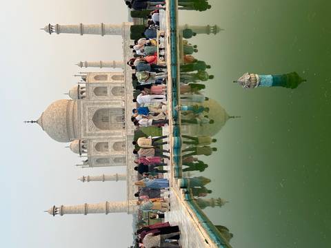 Crowds gather around the reflecting pool in front of the Taj Mahal, whose full facade is mirrored in the green water.