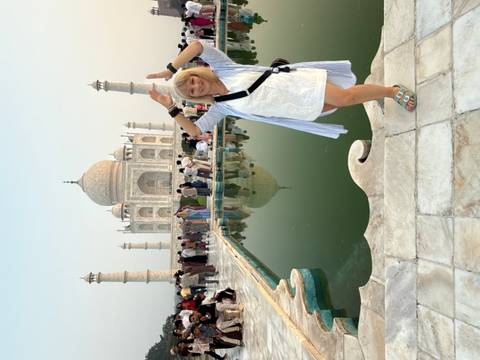 Smiling tourist posing playfully in front of the Taj Mahal reflection pool with crowds behind.