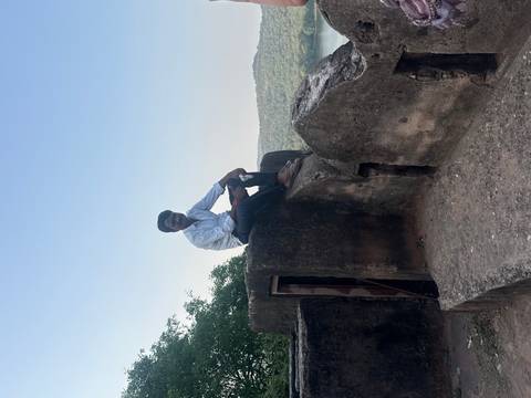 A man sitting on top of an old stone fort wall overlooking green hills under a clear blue sky.