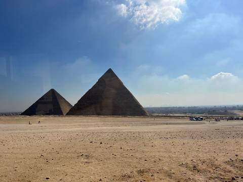 Two of the Giza pyramids dominate a hazy desert plain viewed from a distance under a pale blue sky.