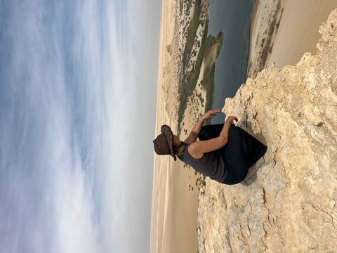 A woman wearing a wide-brim hat crouches on a rocky cliff edge overlooking an oasis and vast desert dunes.