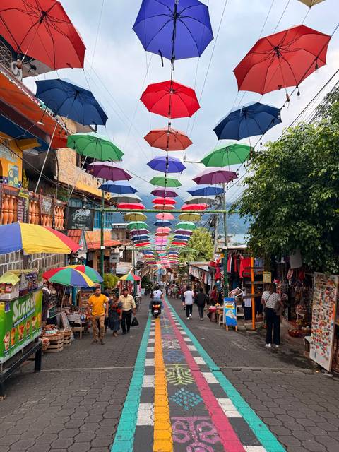 A lively pedestrian lane is covered with colourful umbrellas strung overhead, lined with shops and crowds.