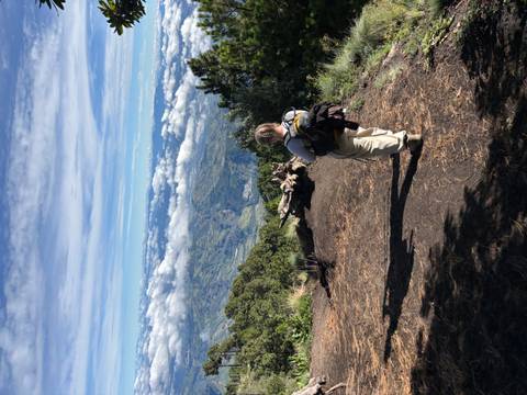 A trekker with a backpack walks along a volcanic ridge with sweeping cloud-filled valleys below.