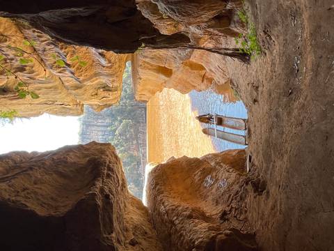Person in a wooden dugout canoe emerging from a sun-lit river canyon framed by towering rock walls.