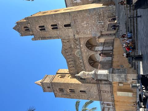 Medieval Cefalù Cathedral with twin towers dominating a sunny stone piazza with tourists sitting nearby.
