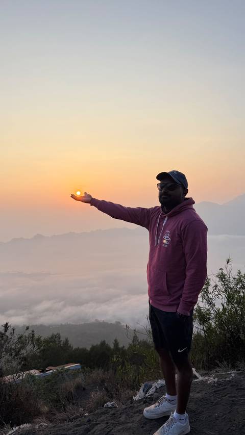 A traveler playfully holds the rising sun in his palm atop a misty mountain viewpoint.