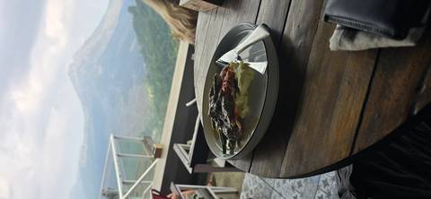 A plated meal on a café table overlooks a hazy volcanic landscape through glass railings.