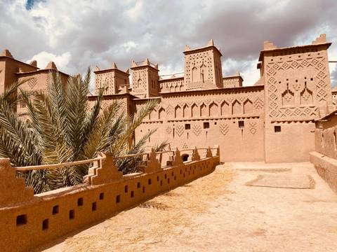 Adobe kasbah with intricate geometric motifs and palm fronds stands under dramatic cloudy skies.