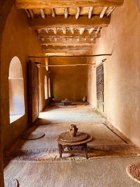 Simple earthen interior room with arched doorways, woven mats and rustic furnishings lit by soft window light.