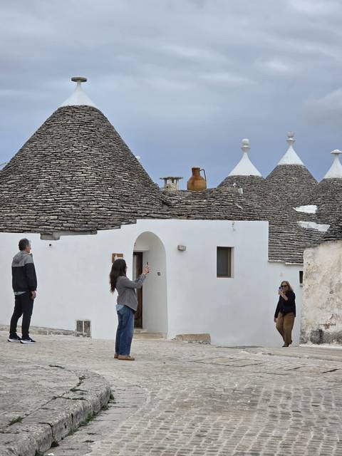 Visitors photograph the whitewashed trulli houses with conical stone roofs in a small lane.