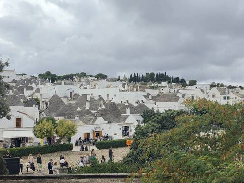 Panoramic view over a hillside town filled with dozens of grey-roofed trulli under a cloudy sky.