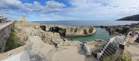Rocky coastal inlet with clear turquoise water, stone steps and a medieval watchtower under a patchy sky.