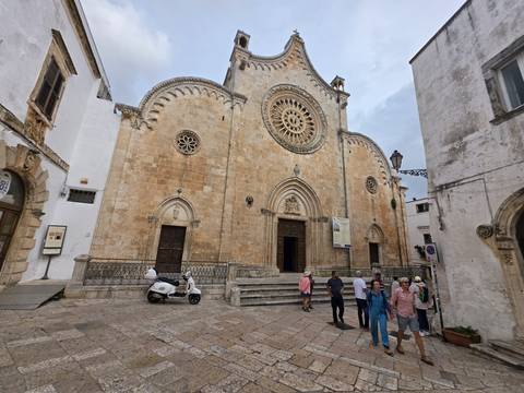 Stone-carved cathedral façade with rose window as tourists gather nearby and a scooter is parked out front.