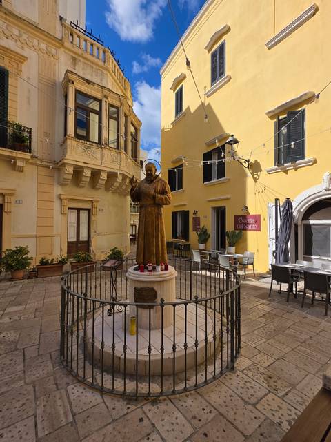 Statue of a saint in a small courtyard surrounded by yellow historic buildings and café tables.