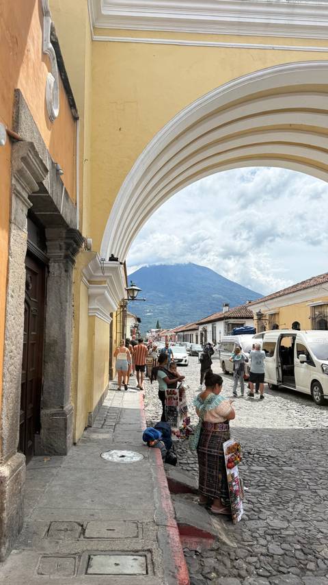 Busy cobblestone street under colonial arches with volcano backdrop and pedestrians.