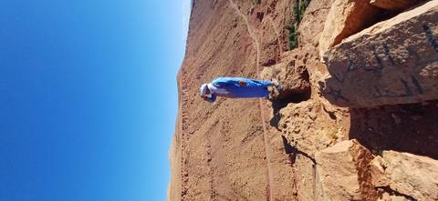 Man in blue robe stands on rocky outcrop overlooking arid red mountains under clear sky.