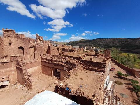 Ruined adobe kasbah walls and towers set against foothill landscape and bright sky.