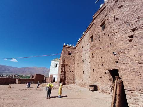 Visitors stand near tall fortress walls built of pinkish stone in a mountain valley.