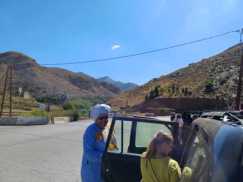 Travel companions prepare to board a 4x4 beside winding mountain road under bright sky.