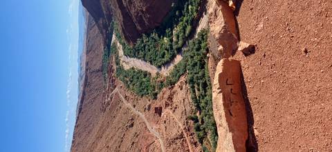 Sweeping desert canyon with ribbon of green oasis and dirt tracks seen from cliff top.