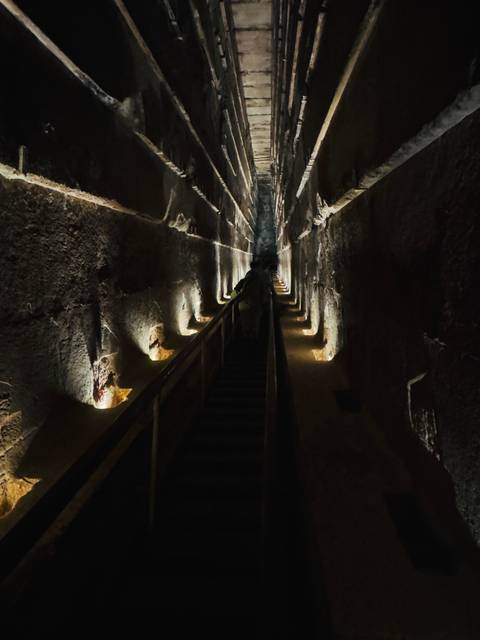 Dimly lit interior corridor of ancient pyramid with steep wooden staircase and stone walls.