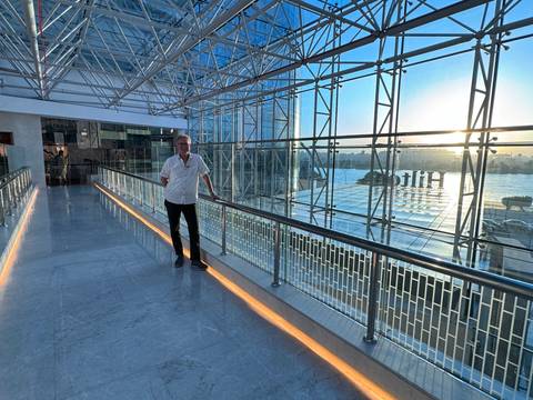 Man stands on glass-roofed walkway overlooking the Nile at sunset inside modern terminal.