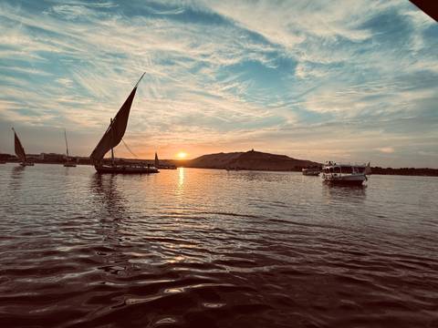 Traditional feluccas sail on the Nile River at fiery sunset with desert hill silhouette.