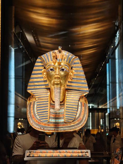 Gold funerary mask of Tutankhamun displayed under spotlights inside a museum gallery.