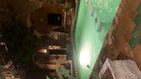 Small indoor pool illuminated at night inside a rustic riad courtyard with earthy walls and plants.