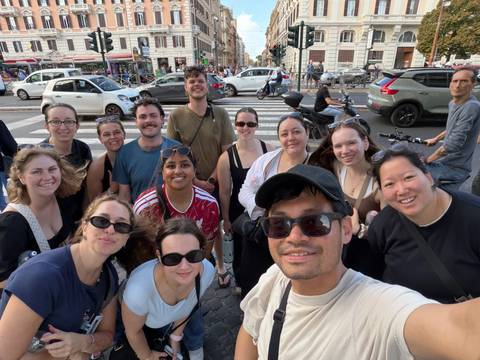 Large group selfie on a bustling European city street with cafes, cars and scooters around.