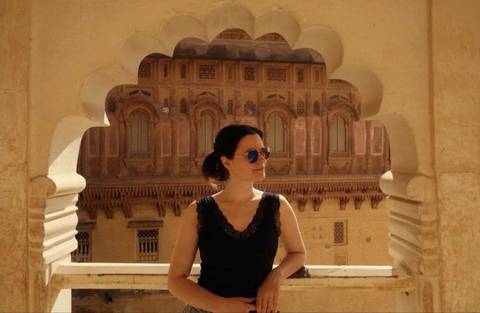 Woman in sunglasses framed by scalloped sandstone arch with ornate palace façade behind.