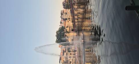 Tall water fountain sprays in a city lake beside a busy arched bridge and colourful buildings.