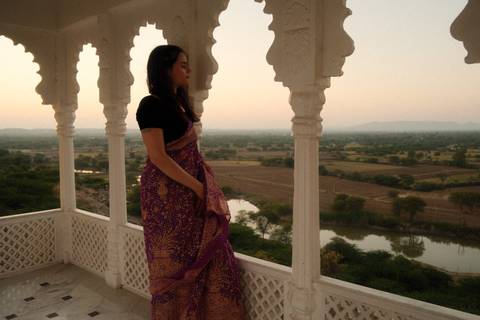 Woman in traditional sari stands on white marble veranda overlooking rural fields at dusk.