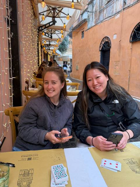 Two smiling women sit at an outdoor cafe table along a narrow European alley decorated with string lights.