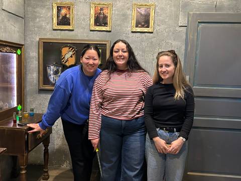 Three women pose together inside a stylish room with framed crests on a textured grey wall.