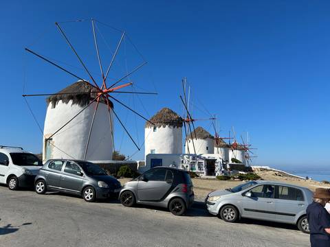 The famous white windmills of Mykonos line the coast behind a row of parked cars under a clear blue sky.